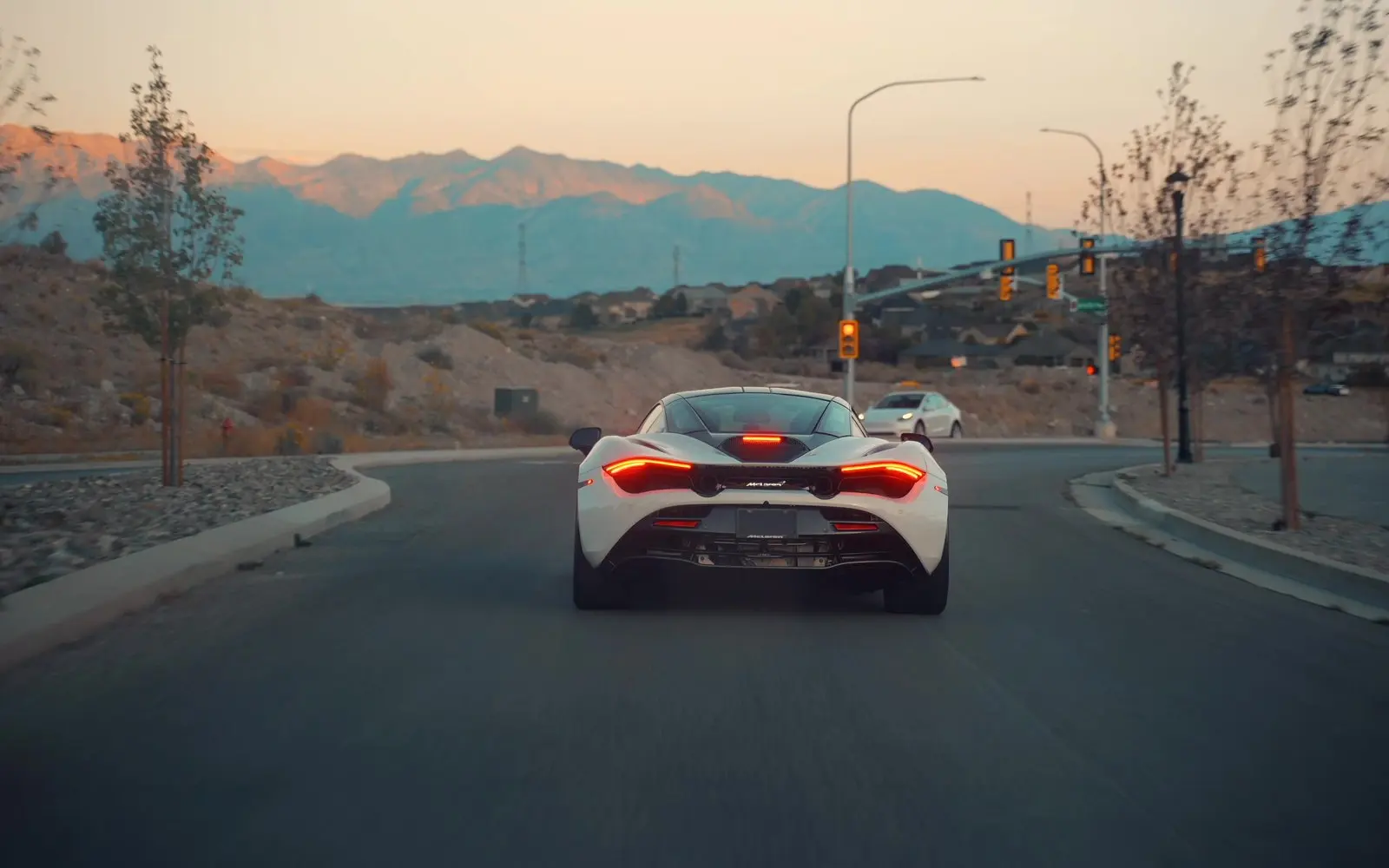 A white McLaren 720S driving away at dusk, taillights lit, mountains ahead