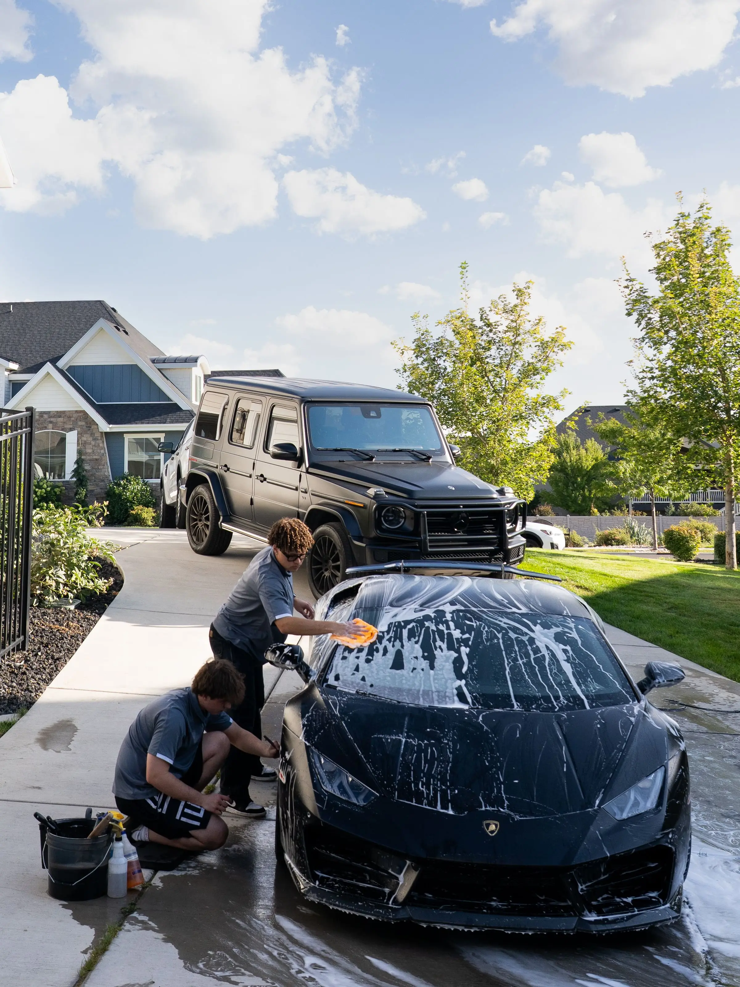 Two detailers washing a foamed black Lamborghini Huracán in a driveway, a Mercedes G-Class parked behind