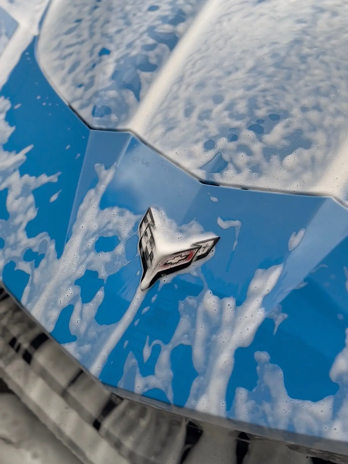 Snow foam on the hood of a blue Corvette C8, sky reflected in the paint