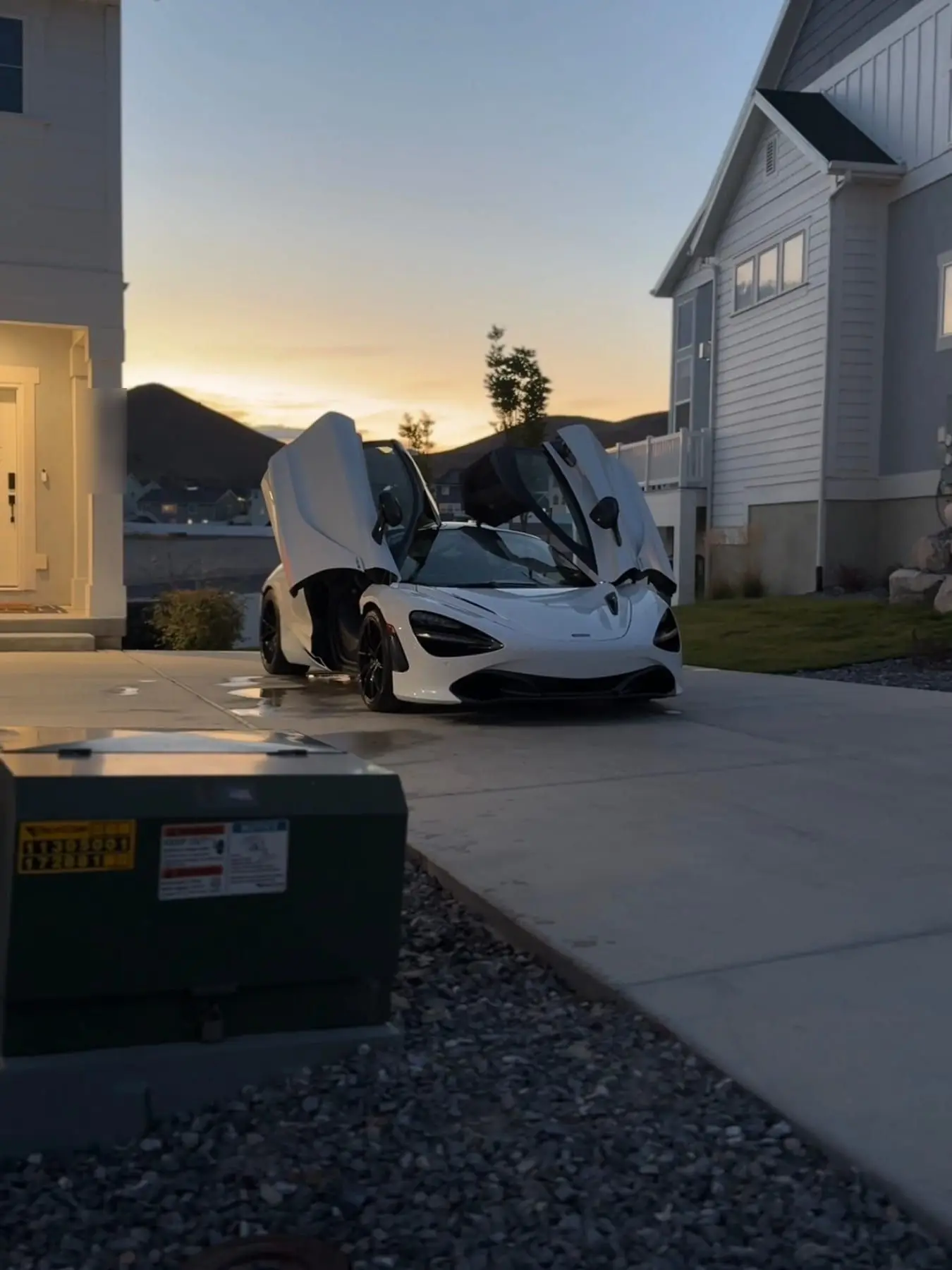 A white McLaren 720S with a door raised in a driveway at sunset